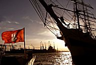 011_15708 Sonnenuntergang im Hamburger Hafen; die Hamburg Fahne strahlt in der Abend Sonne - vom Grosssegler am Kai der Landungsbrcken sind Bug und Masten in der Silhouette zu erkennen - im Hintergrund Krne der Werft an der Elbe.