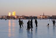 61_5885 Die untergehende Sonne strahlt die Hochhuser an der Mundsburg an - das Licht spiegelt sich auf dem Eis der zugefrorenen Alster. Zwei Schlittschuhlufer spielen Eishockey, andere gehen gegen die Klte dick mit Schal und Mtze eingemummt auf dem Alstereis spazieren. Fotos vom Winter in Hamburg - nach einer langen Frostperiode ist die Auenalster zugefroren.  